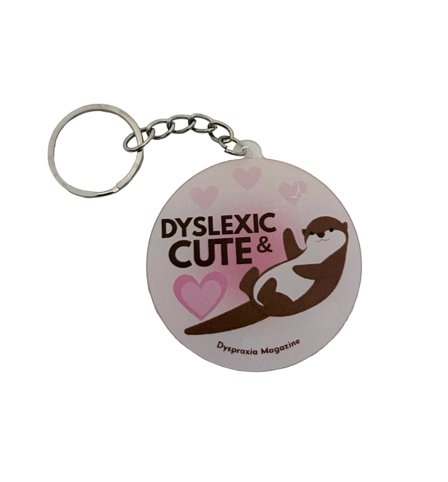 A round acrylic keychain featuring the text 'DYSLEXIC CUTE &' with a cartoon otter and pink hearts, attached to a silver metal keyring on a white textured background.